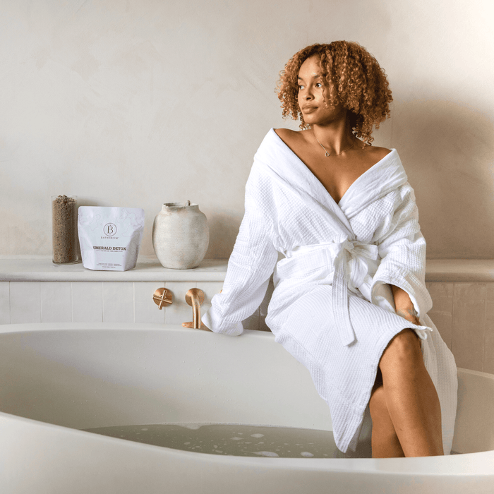 Woman in a white bathrobe sitting on the edge of a bathtub next to a package of Emerald Detox Clay Mineral Soak | Bathorium and spa accessories in a minimalist bathroom setting.