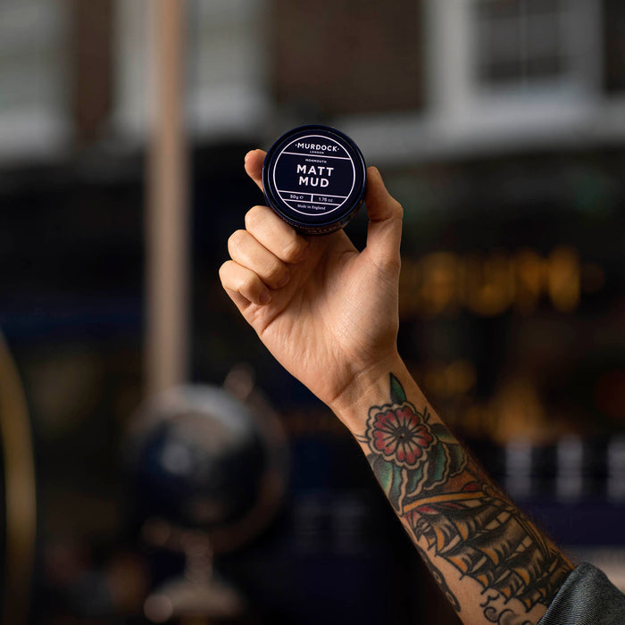 Hånd holder Murdock London matt hårvoks med tatoveringer og barbershop-miljø