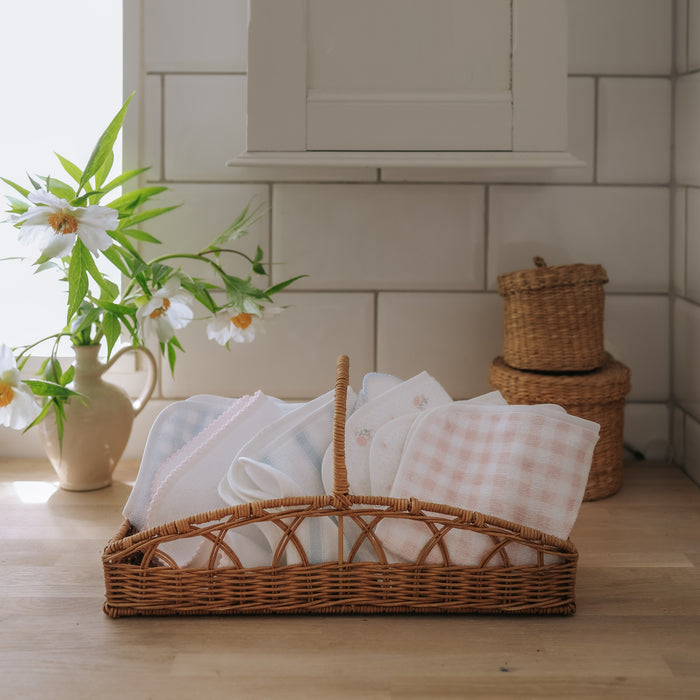 Wicker basket with baby blankets on a wooden floor, with a vase of flowers and woven baskets in the background.