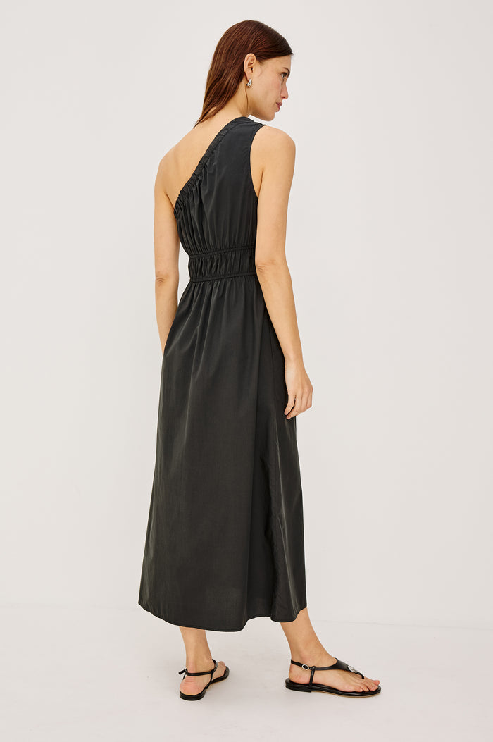 Selani Midi Dress With an Asymmetrical Neckline, Mid-Thigh Slide Slit and Cinched Waistband in Black Colorway - Front Full Body View Featured on Model
