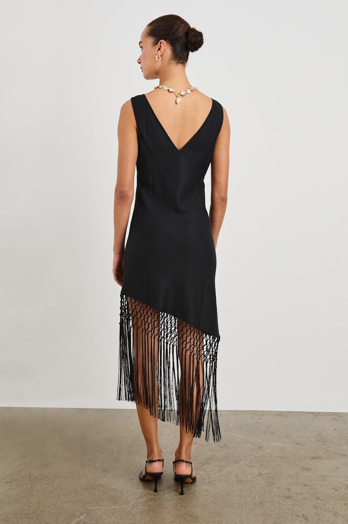 Back view of female model wearing our Tamara Dress in Black. This is a black mini dress with an asymmetrical fringe hem, sheer netted panel, and empire waistline.
