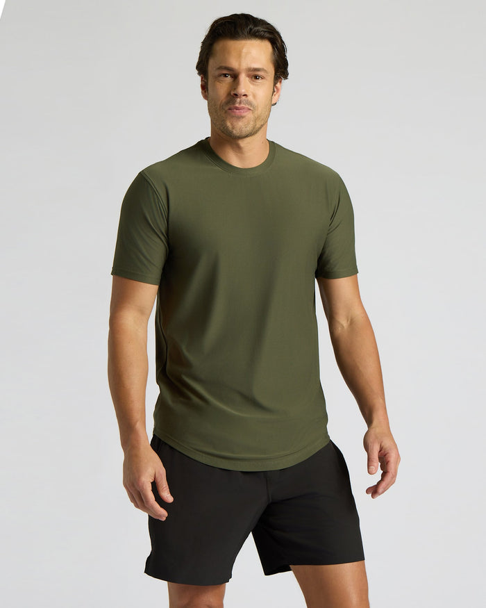 A man wearing a short-sleeve crewneck athletic t-shirt in olive green and black athletic shorts, standing against a light gray studio background.