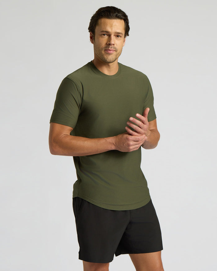 A man wearing an olive green short-sleeve athletic t-shirt and black athletic shorts, standing against a light gray studio background.