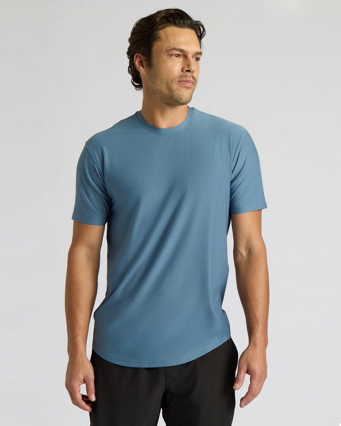 A man wearing a short-sleeve crew-neck athletic t-shirt in a muted slate blue color, paired with black athletic pants, photographed against a neutral light gray studio background.