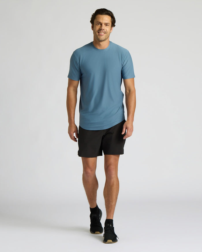 A man modeling a slate blue crewneck t-shirt and black athletic shorts against a light gray studio background.