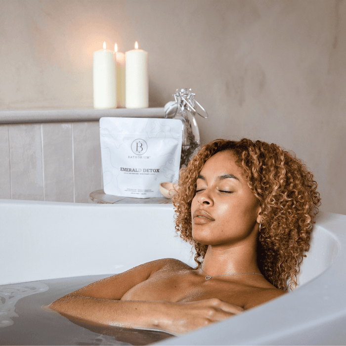 A woman relaxing in a bathtub with eyes closed, next to a package of Emerald Detox Clay Mineral Soak | Bathorium and lit candles creating a calm wellness atmosphere.