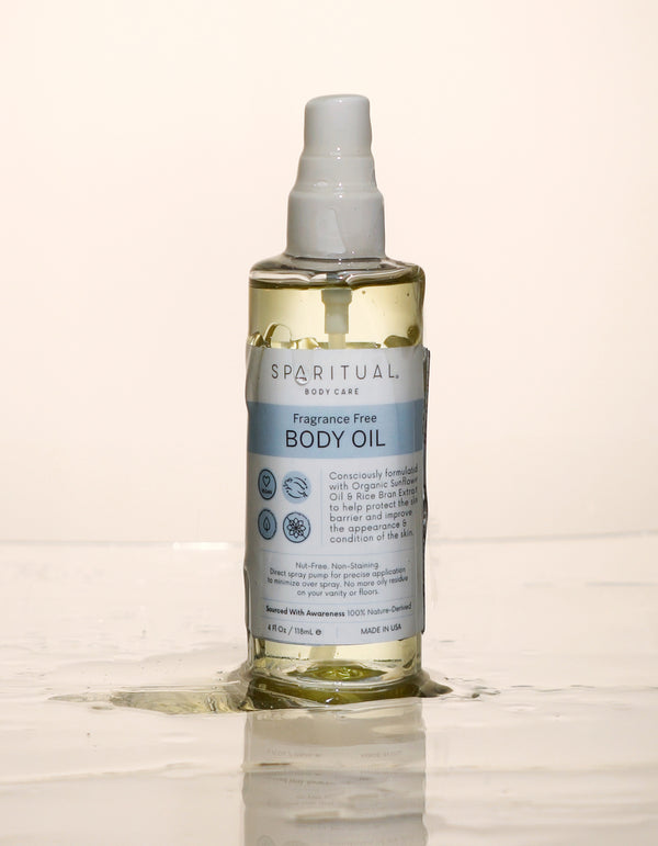 Clear plastic pump bottle of Fragrance Free Body Oil | Sparitual with light yellow oil and blue label standing on wet surface with spilled oil around it, promoting skincare and body care.