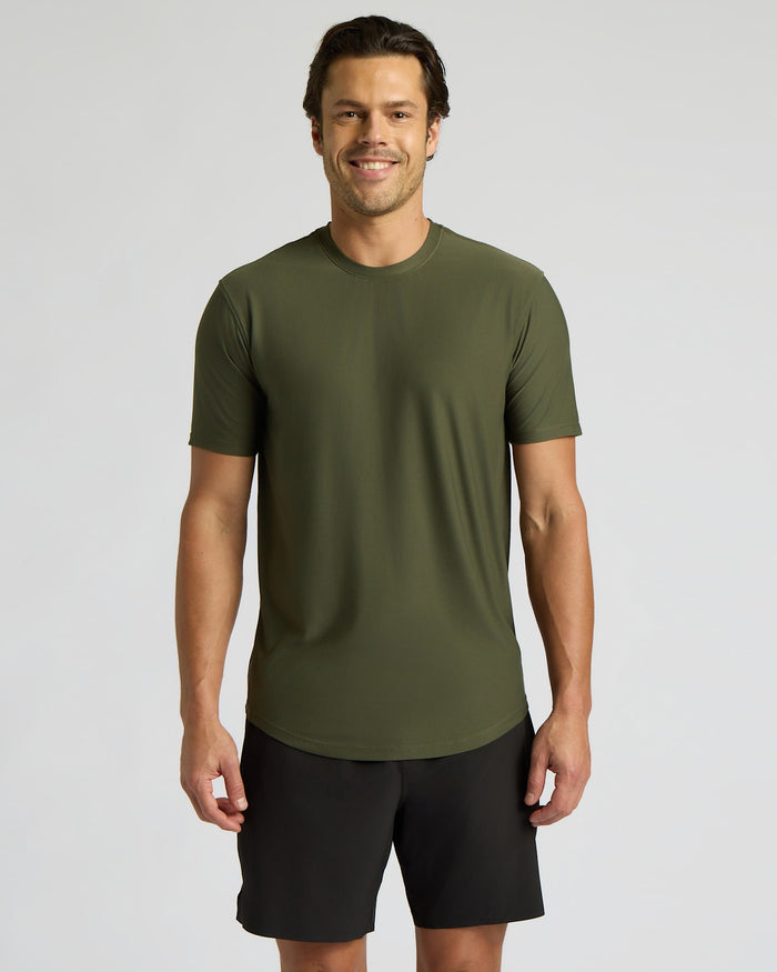 A man wearing a short-sleeve crewneck athletic t-shirt in olive green and black shorts, standing against a light gray studio background.