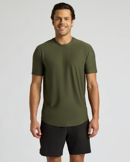 A man wearing a short-sleeve crewneck athletic t-shirt in olive green and black shorts, standing against a light gray studio background.