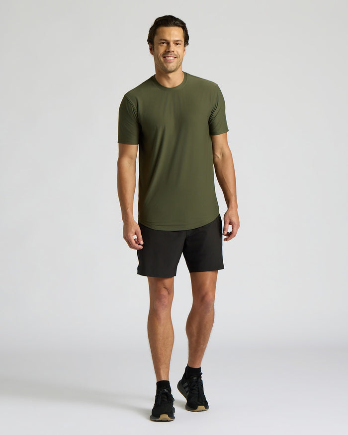 A man modeling a short-sleeve crewneck athletic t-shirt in olive green, paired with black athletic shorts and black running shoes, against a light gray studio background.