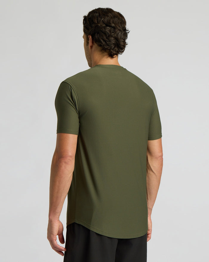 A man viewed from the back wearing a short-sleeve olive green athletic t-shirt and black pants, set against a plain light gray studio background.