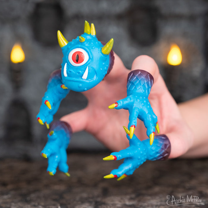 A hand holding a Handimonster, which is a blue one-eyed puppet with yellow horns and sharp claws.