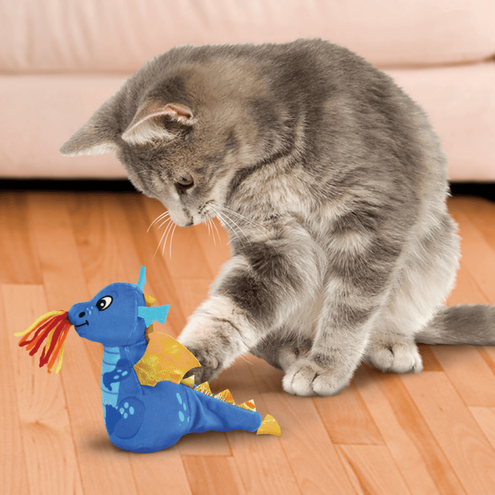 Young grey tabby cat playing with blue plush dragon cat toy with yellow wings and spines, pale blue spots and belly with frame coloured wool threads from it's mouth sat on a laminate floor with a cream sofa in the background.