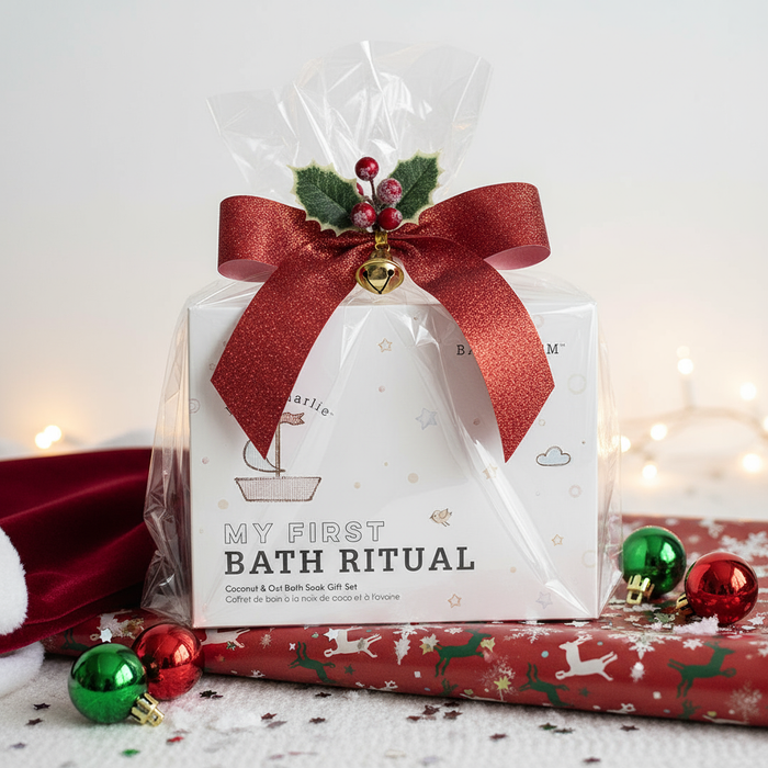 little charlie™ My First Bath Ritual Gift Box Set | Bathorium wrapped in clear plastic with a festive red bow and holly decoration, surrounded by holiday ornaments and lights, perfect for a cozy skincare gift.