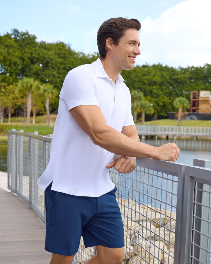 Man in white polo shirt and blue shorts standing on a wooden boardwalk with a scenic background.