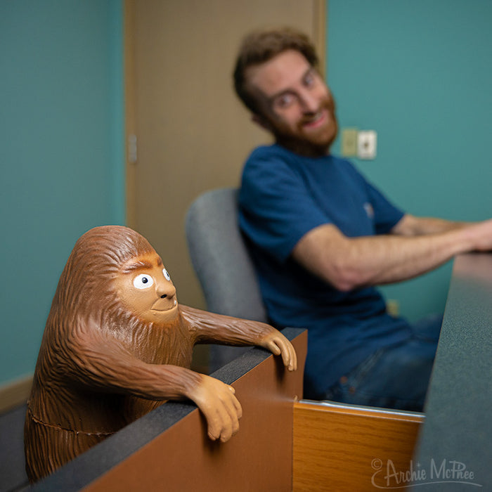 An Office Bigfoot peeks over the desk with a man smiling in the background, sitting in an office setting.