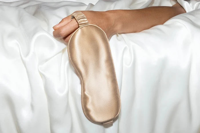 Hand holding a champagne Oi Tape silk sleep mask on white bedding, highlighting soft sheen and contoured shape