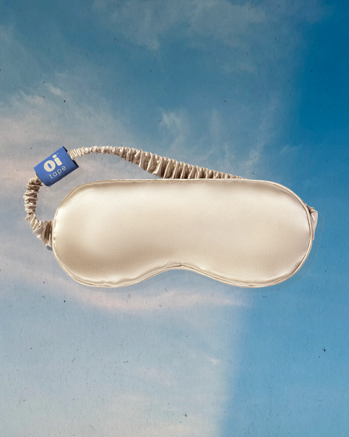 Champagne-colored Oi Tape silk sleep mask on blue sky background, showing ruched elastic strap and smooth finish.