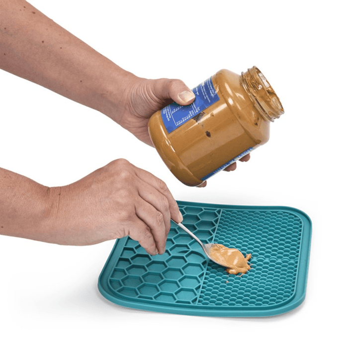 One hand holding a jar of pet safe nut butter and the other hand spooning the butter onto a blue silicone lick mat with two different textures, one with hexagon shapes, the other with small close together ridges pictured on a white background.