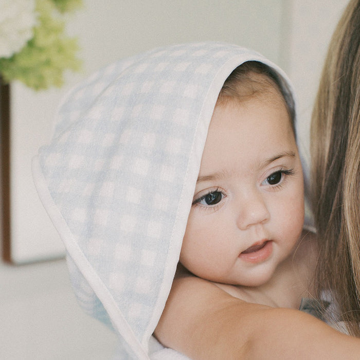 A close-up of a baby wrapped in a light blue and white gingham checkered towel, held by an adult whose arm is visible on the right.