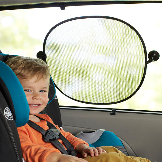 A young child smiles in a car seat beside a Nuby Pop Open Car Sunshade (3 Pack), which blocks sunlight and glare. The car’s interior is visible, with greenery outside the window.