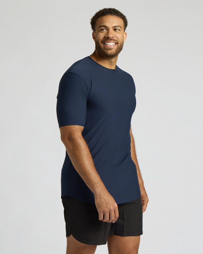 Male model wearing a navy blue Tall Active Curved Hem Crew 6-Pack shirt paired with black athletic shorts, smiling and looking off to the side against a plain background