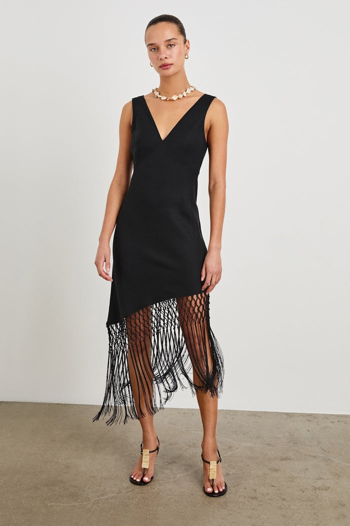 Front view of female model wearing our Tamara Dress in Black.  This is a black mini dress with an asymmetrical fringe hem, sheer netted panel, and empire waistline.