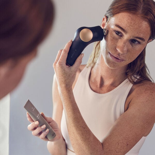 Woman using a facial wellness device on her cheek while holding a tube of TheraFace Conductive Gel | Therabody for skincare and body care routine.