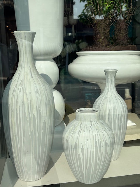 A set of white and gray matte finish vases of various sizes displayed in a storefront window, with one vase containing a green plant.